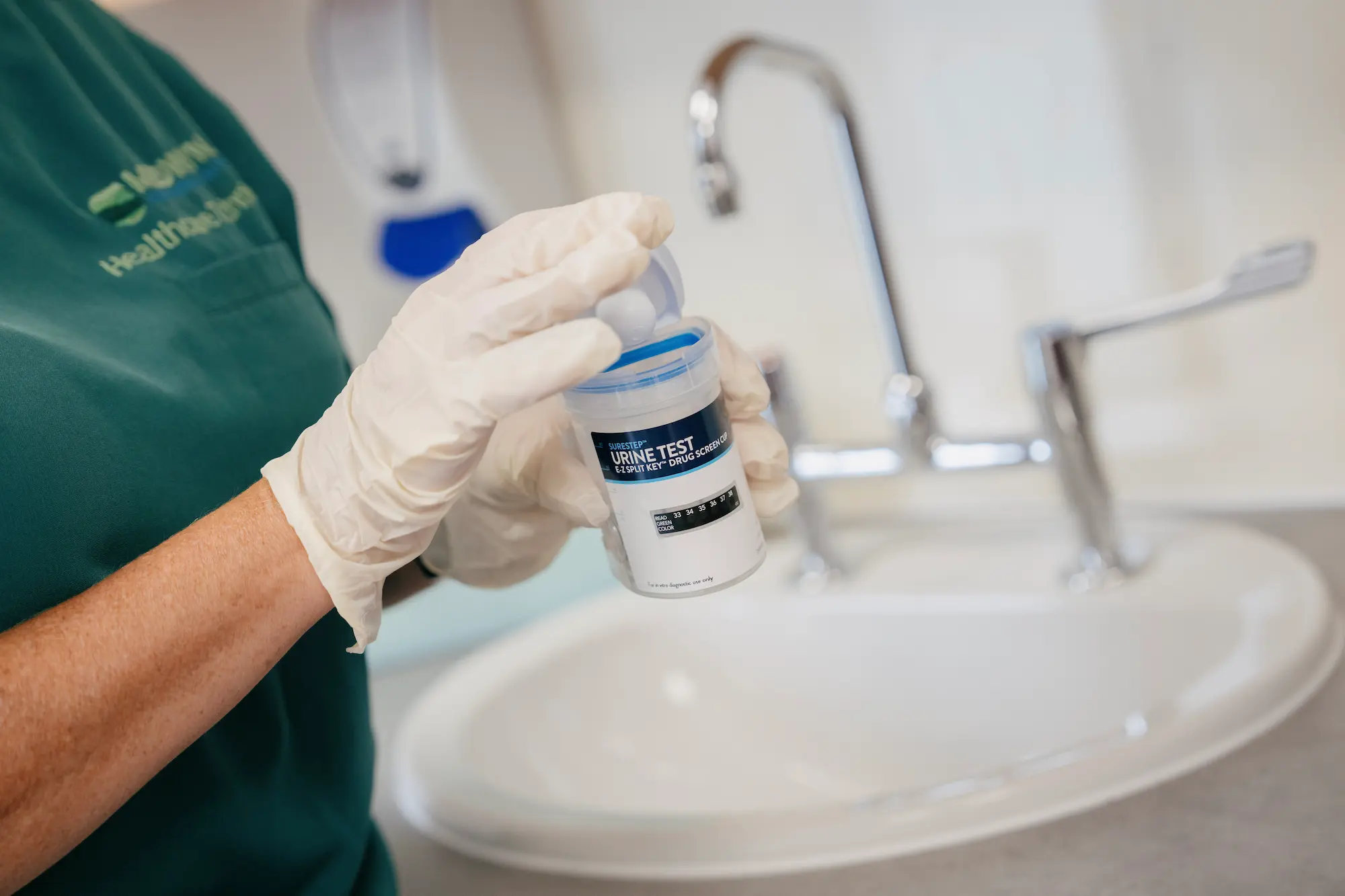 A gloved Medwyn healthcare technician holding a sealed urine drug screen cup during a workplace drug test